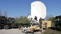 Mehrere Soldaten klappen eine Antenne für satellitenkommunikation auf, die auf einem Anhäger steht. 