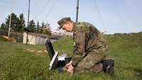 Ein Soldat kniet auf einer Wiese und baut eine Funkantenne auf.
