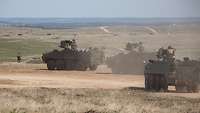 Vier Radpanzer fahren über weites, trockenes Gelände, Sand wirbelt auf.