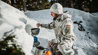 Ein Mann sitzt im Schnee und rührt in einem Topf auf dem Gaskocher