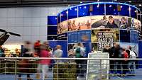 Ein Messestand mit blauem Rundbau ist zu erkennen, an Ständen drumherum befinden sich Besucher und Soldaten.