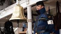 Ein in blau gekleideter Soldat steht vor einer goldenen Glocke mit er Aufschrift „Gorch Fock".