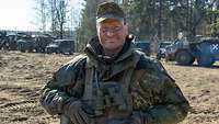 Ein Soldat der Bundeswehr lächelt für eine Portraitaufnahme in die Kamera, im Hintergrund ist ein Gefechtsstand erkennbar.