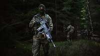 Ein bewaffneter Soldat steht im Tannenwald bei Abenddämmerung. Im Hintergrund weitere Soldaten.
