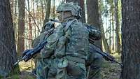 Mehrere Soldaten der Bundeswehr hocken in Stellung zwischen dicht beieinander stehenden Bäumen in einem Wald.