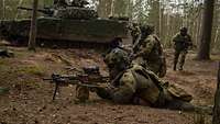 Soldaten legen sich auf Waldboden in Stellung, dahinter ist ein Fahrzeug vom Typ Schützenpanzer Combat Vehicle 90.