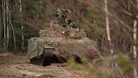 Auf sandigen Waldboden steht ein Fahrzeug vom Typ Combat Vehicle 90 zwischen Bäumen.