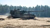Eine Panzerhaubitze 2000 fährt über offenes sandiges Gelände, dahinter ist eine Waldkette zu erkennen.