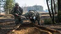Zwei Soldaten arbeiten mit Schaufel und Eimer an einem Schützengraben in einem Wald.