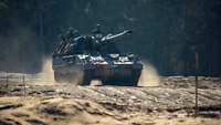 Mehrere Soldaten fahren auf einer Panzerhaubitze 2000 durch offenes Gelände, im Hintergrund erkennt man Bäume.