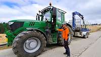Ein Mann mit orangener Jacke hält die Tür eines grünen Traktor mit Anhänger offen