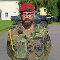 Ein Soldat mit rotem Barett, Brille und Bart schaut freundlich in die Kamera.