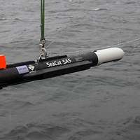 Eine Metallröhre in den Farben Grau, Schwarz und Orange#en hängt über Wasser an einem Haken.