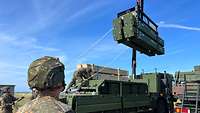 Ein Kran hebt vier schwere Metallblöcke auf ein Militärfahrzeug.
