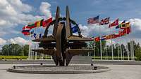 Die NATO-Sternenskulptur und dahinter wehen die Flaggen der Mitgliedsstaaten an Flaggenmasten