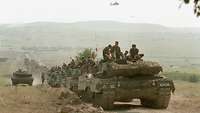 KFOR-Truppen im Kosovo Panzer mit Deutschlandflaggen fahren im Konvoi. Zwei Hubschrauber fliegen in der Luft.