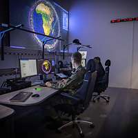 Zwei Personen sitzen in einem großen Raum mit vielen Bildschirmen und Computern.