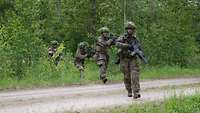 Drei bewaffnete Soldaten überqueren hintereinander eine Straße im Wald. 