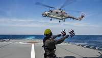 Ein Fluglotse steht am dem Heck eines Marineschiffes und weist einen landenden Hubschrauber ein.
