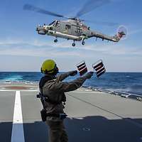 Ein Fluglotse steht am dem Heck eines Marineschiffes und weist einen landenden Hubschrauber ein.