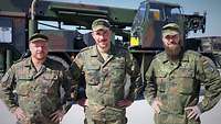 Drei Soldaten stehen nebeneinander vor einem Kranfahrzeug.