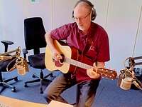 Militärpfarrer Jens Augustin spielt Gitarre Ein Mensch hat Kopfhörer auf, sitzt auf einem Stuhl und spielt Gitarre.