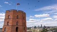 Über einem Backsteinturm mit Nationalflagge eine Flugformation, im Hintergrund das Stadtpanorama.
