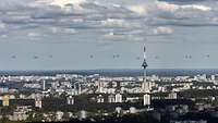 Blick über eine Stadt mit Hochhäusern und einem Fernsehturm fliegen viele Hubschrauber.