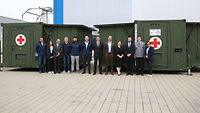 Mehrere Menschen stehen nebeneinander vor zwei dunkelgrünen Container.