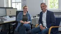 Eine Frau und ein Mann sitzen ein einem Büro und blicken in die Kamera