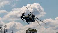 Ein Hubschrauber fliegt mit hoher Geschwindigkeit eine Kurve über Bäumen bei blauem Himmel.