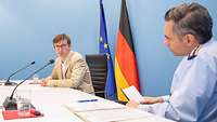 Ein Soldat und ein Mann sitzen an einem Tisch während eines Interviews. Im Hintergrund zwei Flaggen.