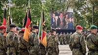 Ein Platz umsäumt mit angetretenen Soldaten, ein Politiker ist auf einer Leinwand zu sehen.