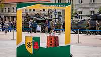 Panzer stehen am Straßenrand, davor ein großer grün-gelber Aufsteller mit Wappen.