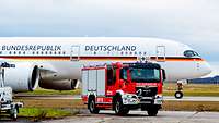 Ein Feuerwehrfahrzeug steht auf einem Flugplatz vor einem Passagierflieger der Bundesrepublik