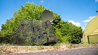 Eine große olivgrüne Satellitenschüssel mit zugehöriger Technik steht abgetarnt vor einem Gebüsch.