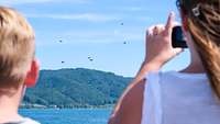 Two onlookers standing in the foreground watch parachutists above a lake
