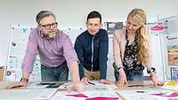Ein Mann, ein junger Erwachsener und eine Frau stehen vor einem Tisch und schauen sich Dokumente an