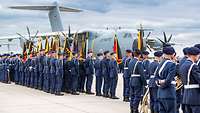 Soldaten stehen in Formation mit Truppenfahnen auf einem Platz, im Hintergrund steht ein Flugzeug