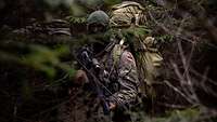 Ein vermummter Soldat steht in einem Wald mit einem großen Rucksack auf dem Rücken.