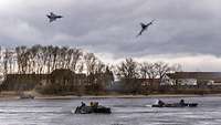 Zwei Kampfflugzeuge fliegen am Himmel, während zwei Transportpanzer durch einen Fluss fahren.