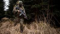 Ein vermummter Soldat geht in einem Wald mit einem großen Rucksack auf dem Rücken.