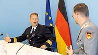 Ein Soldat interviewt einen anderen Soldaten sitzend an einem Tisch. Im Hintergrund stehen Flaggen.