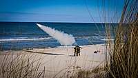Zwei Soldaten stehen am Strand und schießen eine Flugabwehrrakete in den Himmel über das Meer.