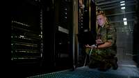 Ein uniformierter Soldat kniet mit einem Laptop in einem Serverraum.