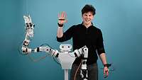 Ein junger Mann winkt mit dem rechten Arm, vor ihm steht ein weißer Roboter