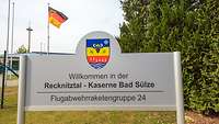 Ein Willkommensschild mit Wappen und Grußworten steht vor einem Zaun und einer Deutschlandfahne.
