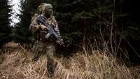 Ein vermummter Soldat mit einem Rucksack und Waffe geht durch waldiges Gelände