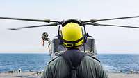 Ein Soldat mit einem gelben Helm steht vor einem Helikopter, der auf einem Schiff gelandet ist.