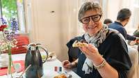 Eine Frau lacht in die Kamera während sie frühstückt.